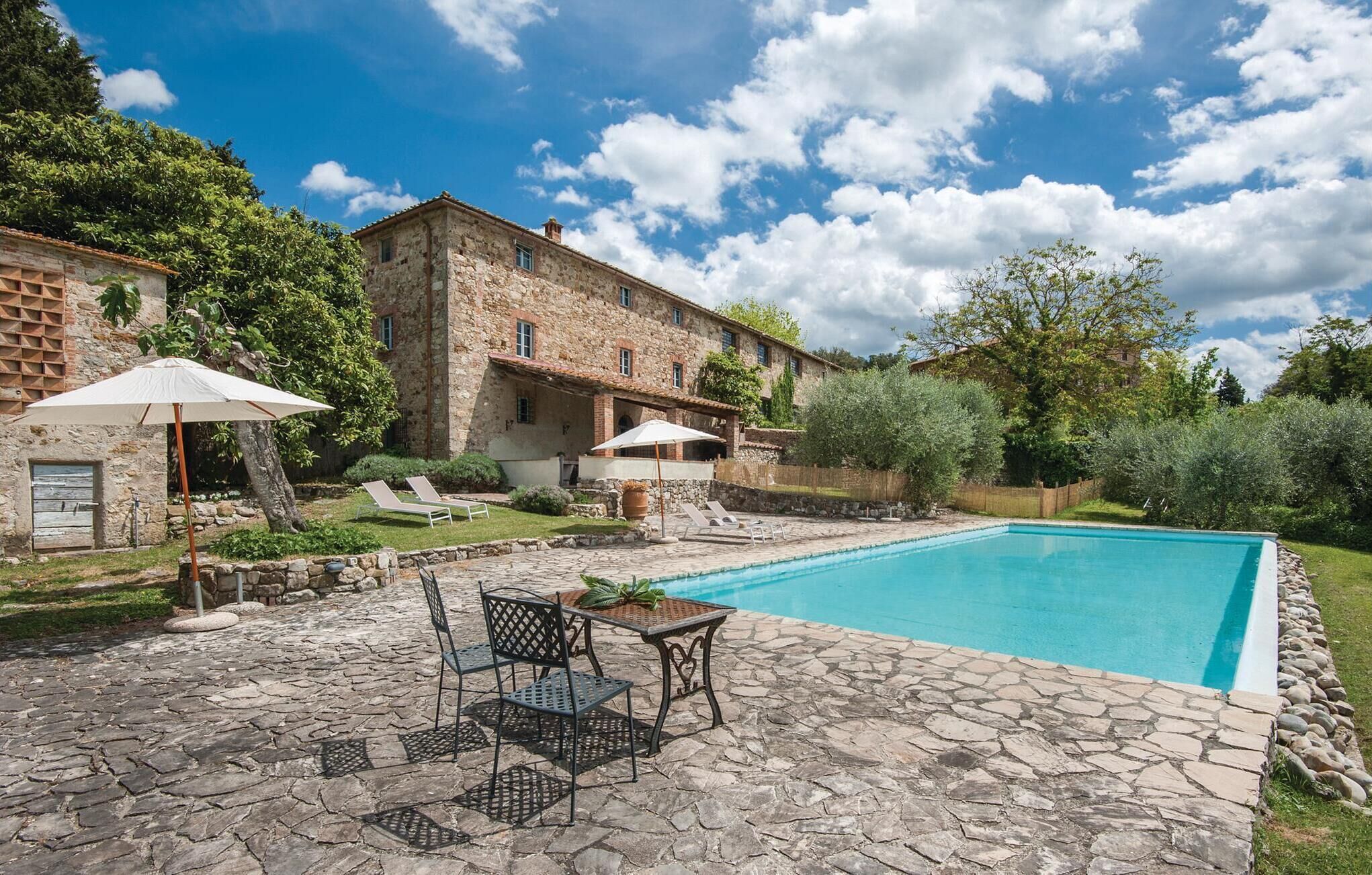 A stone pool with lounge chairs and umbrellas, surrounded by a stone patio and a stone building with a garden.