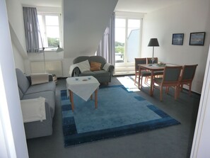 Ein Wohnzimmer mit einer Couch, einem Tisch, Stühlen und einem Fenster mit Vorhängen, sowie einem Blick ins Freie.