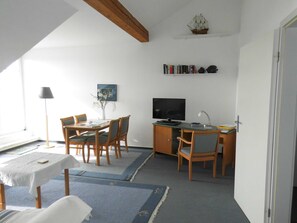 Ein Zimmer mit Esstisch, Stühlen, einem Fernseher, einem Bücherregal und einem Schiffsmodell an der Wand.