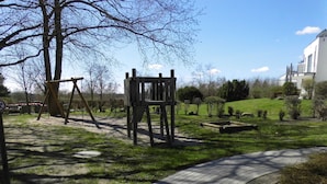 A park with a swing set, a wooden climbing structure, a paved path, and a grassy area with trees and shrubs.