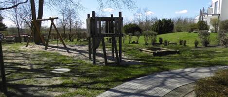 Un parc équipé d’une balançoire, d’une structure d’escalade en bois, d’un chemin pavé et d’une zone herbeuse agrémentée d’arbres et d’arbustes.