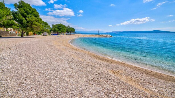 A pebble beach with clear blue water, trees, and a distant shoreline. - Holiday apartment with balcony and air conditioning (Crikvenica)
