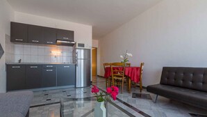 Eine moderne Küche mit dunklen Schränken, einem Edelstahlkühlschrank und einer Mikrowelle. Ein Essbereich mit roter Tischdecke und Stühlen sowie ein Sofa im Wohnbereich.