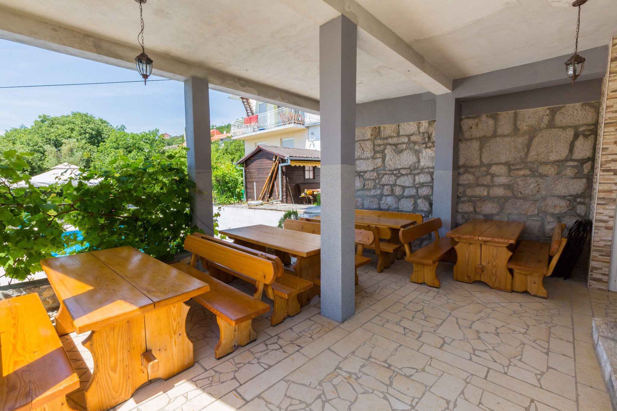 An outdoor patio area with wooden tables and benches, stone walls, and a view of a residential building in the background.