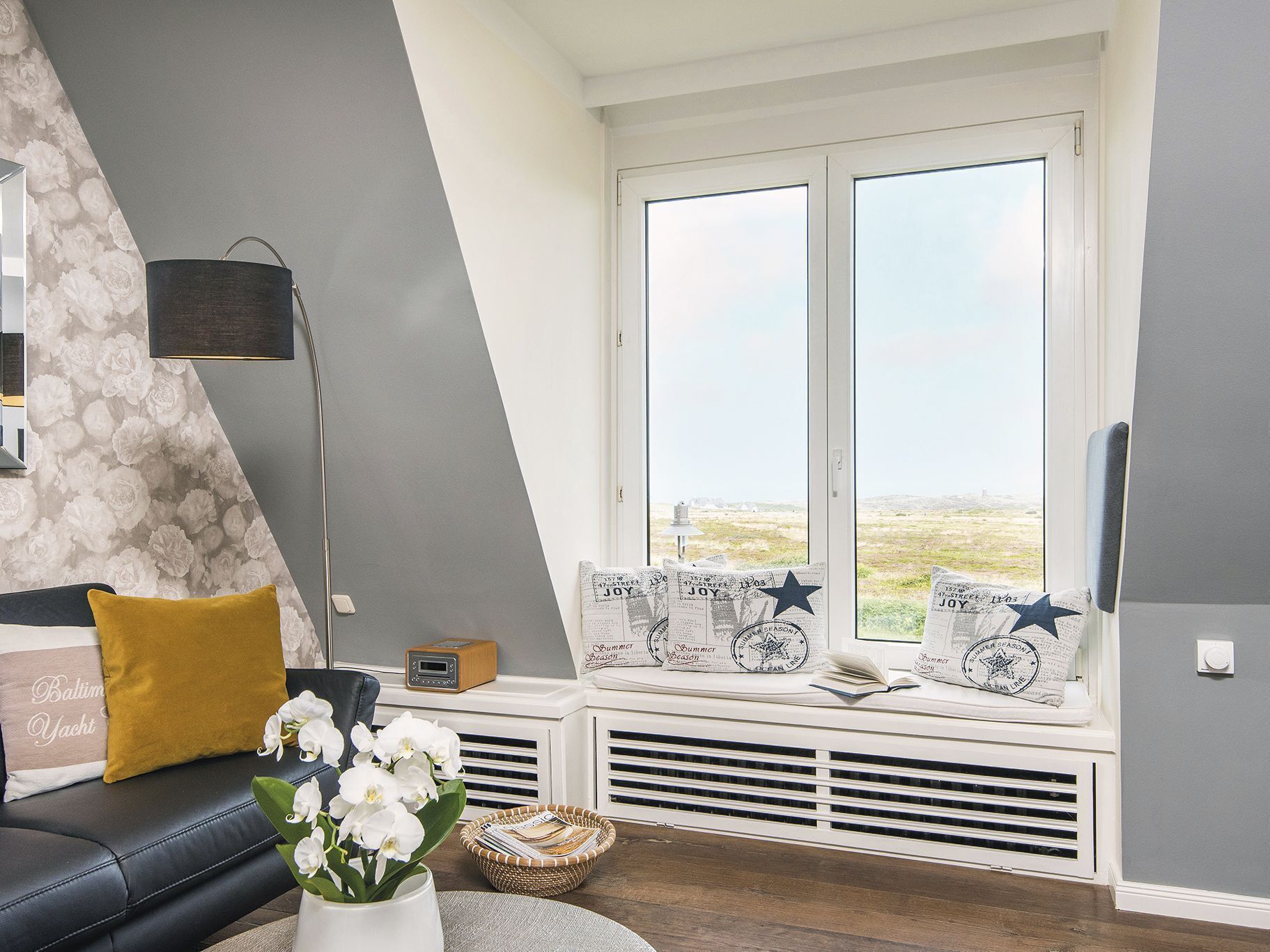 Un salon moderne avec un canapé en cuir noir, un coussin de couleur moutarde, un vase blanc orné de fleurs et une fenêtre donnant sur la campagne.