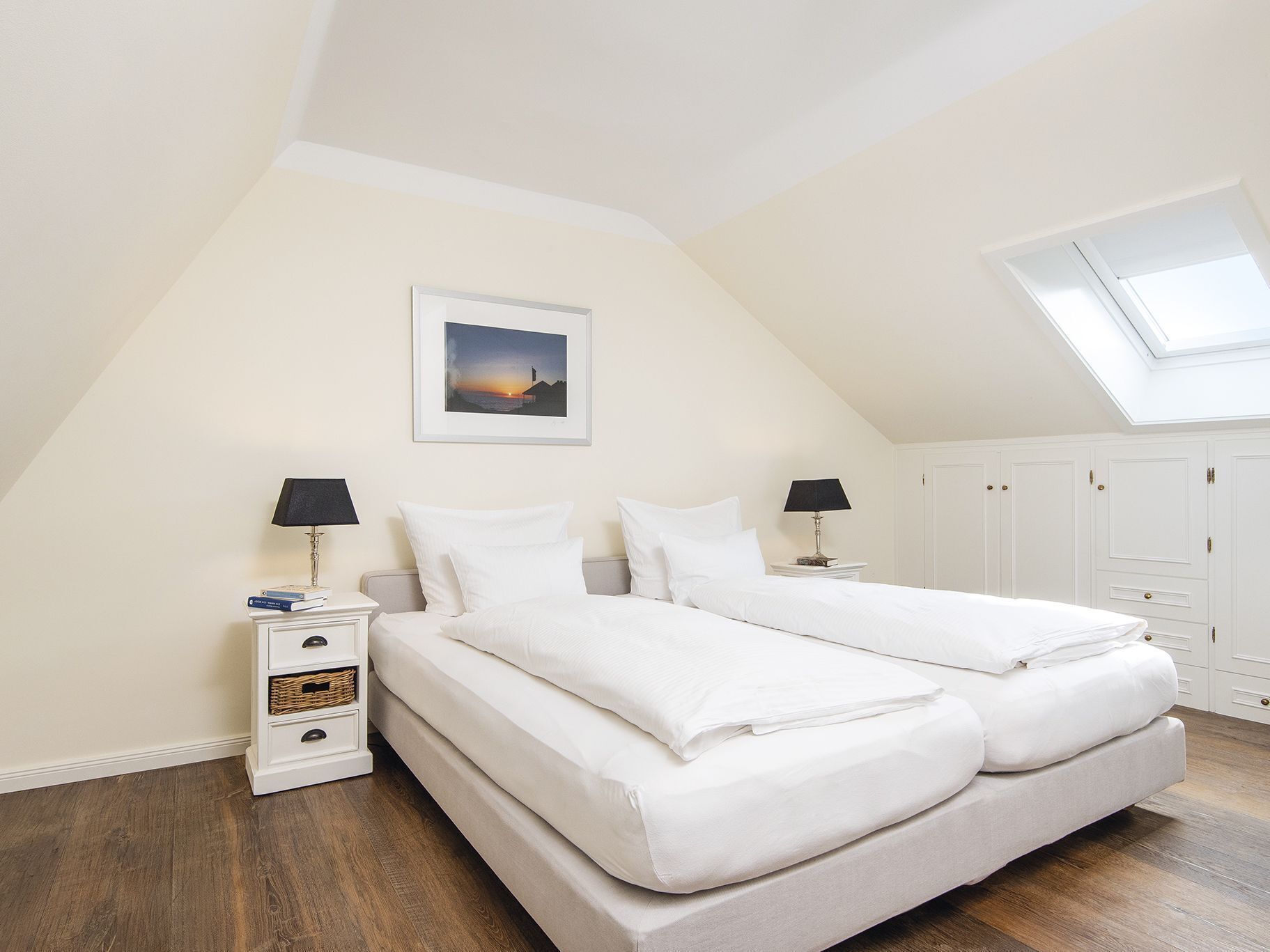 A bedroom with a skylight, a bed with white linens, a nightstand with a lamp, and a framed picture on the wall.