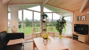 Una sala de estar acogedora con un sofá de cuero negro, una mesa de centro de madera con un jarrón de flores, un televisor sobre un mueble de madera y ventanas amplias con vistas al exterior.