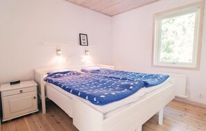 A bedroom with a white bed, a nightstand, a wall-mounted light, a window with a view of greenery, and a framed picture above the bed. - 4 bedroom stunning home in Slite (Slite)