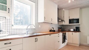 Cocina moderna con armarios blancos, encimera de madera, microondas negro y ventana con vistas a árboles.