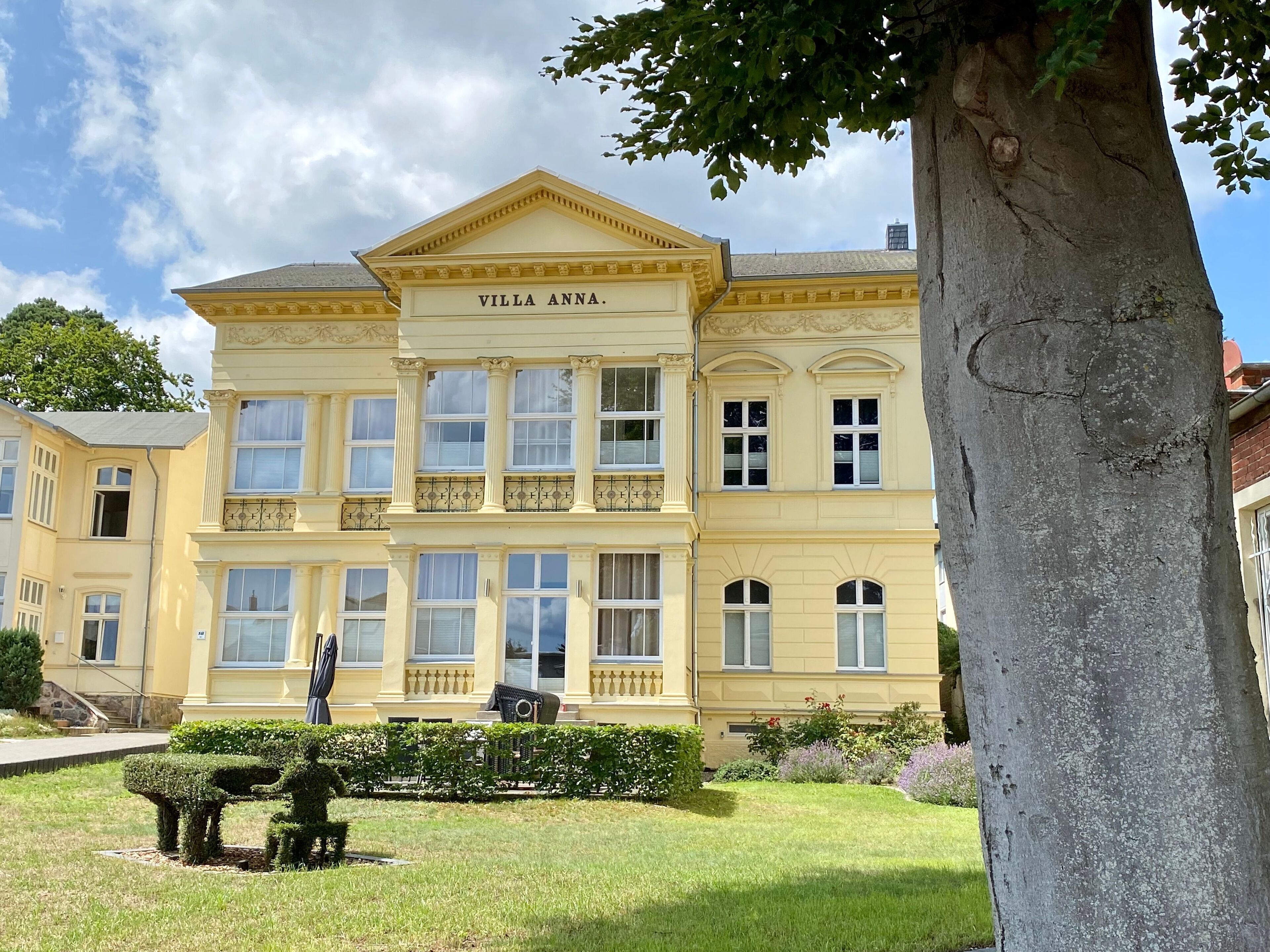 Ein großes, gelbes Gebäude mit der Aufschrift 'Villa Anna' an der Fassade, umgeben von gepflegten Rasenflächen und einem Baum.
