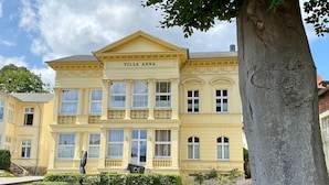 Een groot, geel gebouw met de naam 'Villa Anna' op de gevel, omgeven door keurig onderhouden gazons en een boom.