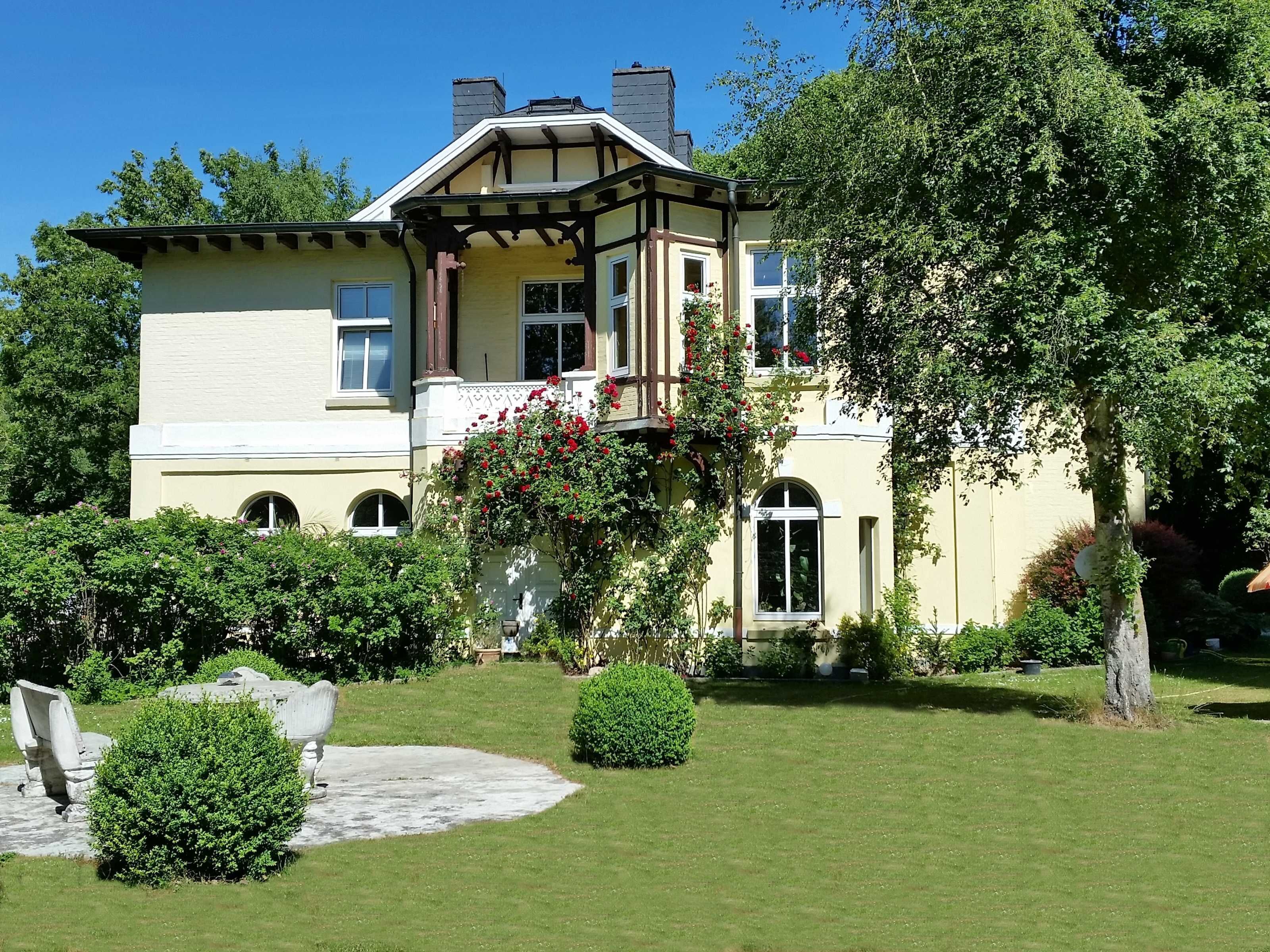 Ein großes, zweistöckiges Haus mit einem Balkon, umgeben von gepflegten Gärten und Bäumen.