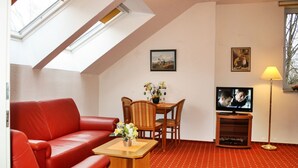 A living room with a red sofa, a wooden table with a vase of flowers, a television on a stand, and a red patterned carpet. - 2-room apartment no. 6, 35m², DG, West - Waldhaus Gronenberg (Scharbeutz)