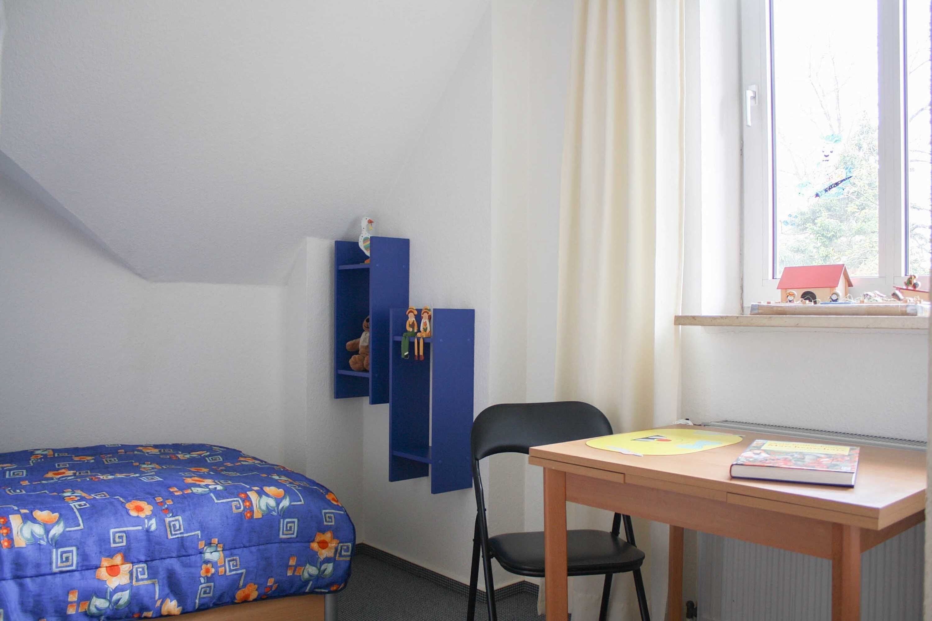 Huoneessa on sänky, työpöytä, tuoli, ikkuna, jossa on verhot, ja hylly, jossa on leluja.
