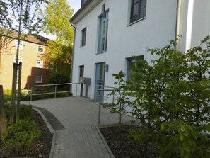 Un bâtiment avec une entrée pavée, une porte bleue et un petit arbre au premier plan.