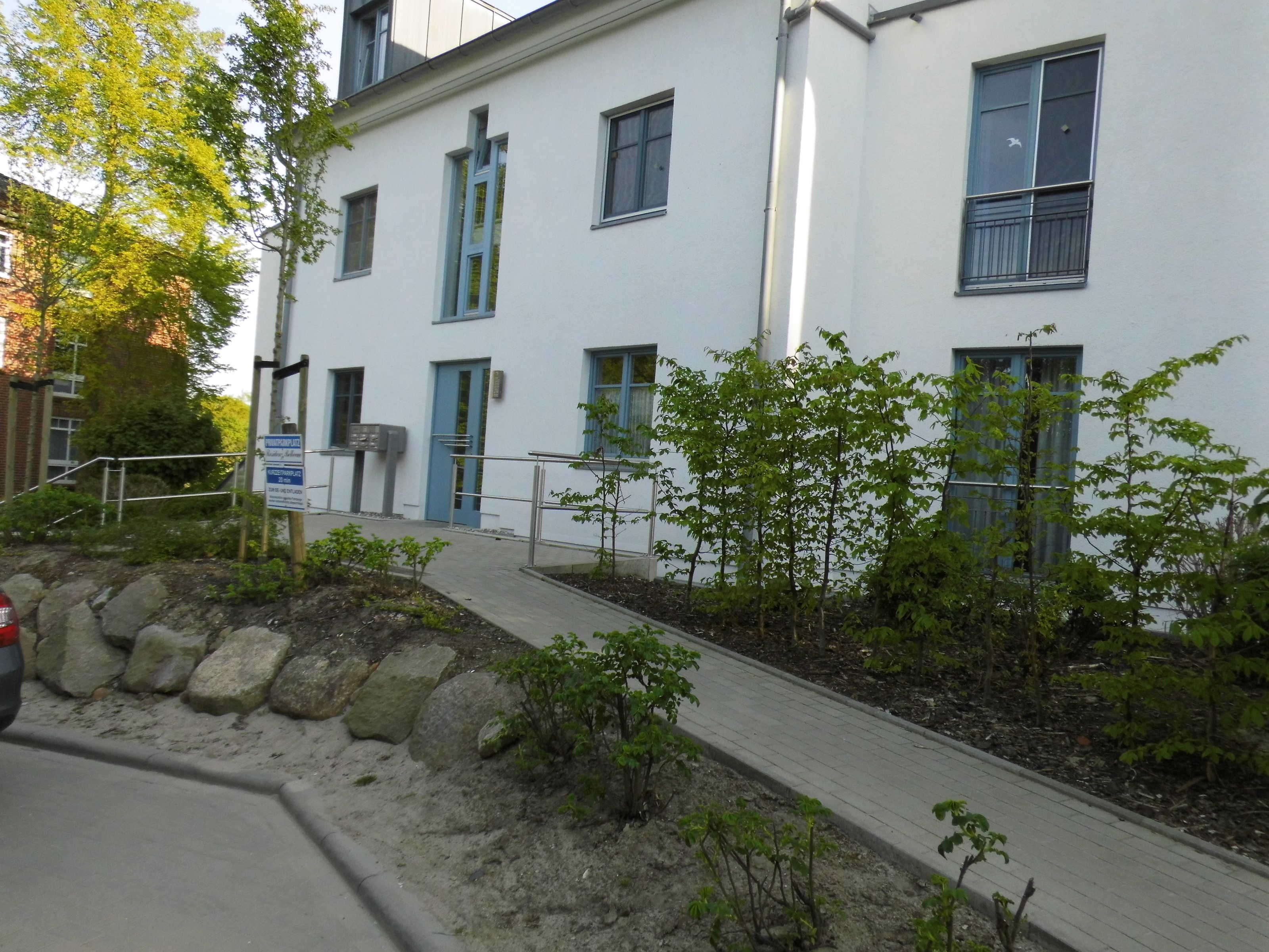 A building with multiple windows, a paved pathway, and a small garden area with young trees.