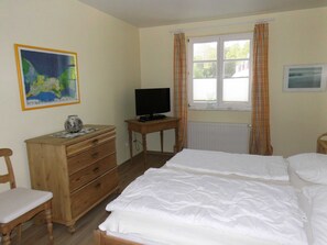 Ein Schlafzimmer mit Bett, einem Holzkapfenkasten, einem Fernseher auf einem kleinen Tisch und einem Fenster mit Vorhängen.