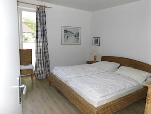 Ein Schlafzimmer mit einem Doppelbett, einem Korbstuhl, einem Nachttisch aus Holz mit Lampe und einem Fenster mit Vorhang.