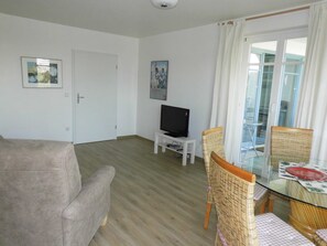Ein Wohnzimmer mit einer Couch, einem Fernseher auf einem Schrank, Korbstühlen, einem Glastisch und einer Tür.