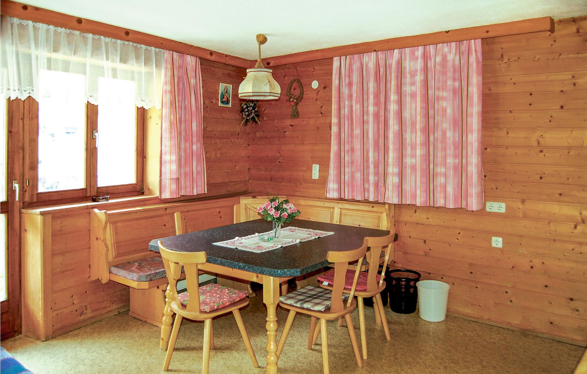 A wooden cabin interior with a dining table, four chairs, a vase of flowers, and a hanging light fixture.