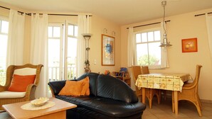 A living room with a black leather sofa, a wooden coffee table, wicker chairs, a dining table with a floral tablecloth, and large windows with curtains. - Amber Whg. BE-10 .. - Amber Whg. 10 (Kühlungsborn)