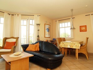 Un salon avec un canapé en cuir noir, une table basse en bois, des fauteuils en osier, une table à manger recouverte d’une nappe à motifs floraux et de grandes fenêtres avec des rideaux.