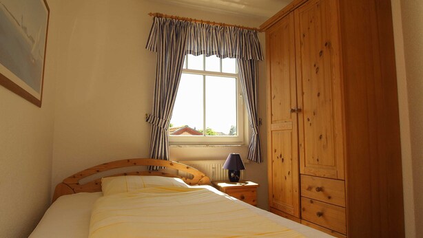 A bedroom with a bed, a wooden wardrobe, a window with curtains, and a lamp on a bedside table. - Amber Whg. BE-10 .. - Amber Whg. 10 (Kühlungsborn)