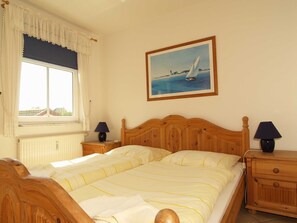 Un lit double avec une tête de lit en bois, deux tables de chevet avec des lampes, une fenêtre avec des rideaux et un tableau représentant un voilier accroché au mur.
