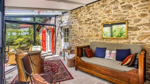 A cozy room with a stone wall, a wooden sofa with cushions, wicker chairs, and a red patterned rug.