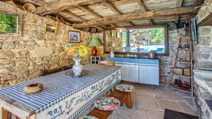 A rustic stone-walled room with a wooden ceiling, a table with a checkered tablecloth, a vase with flowers, and a mirror reflecting an outdoor pool area.