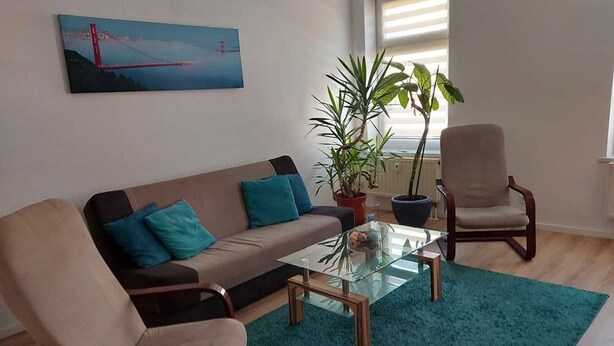 A living room with a sofa, two armchairs, a glass coffee table, a potted plant, and a painting of a bridge. - Vacation apartments in Chemnitz - Holiday apartment 1 Red (Chemnitz)