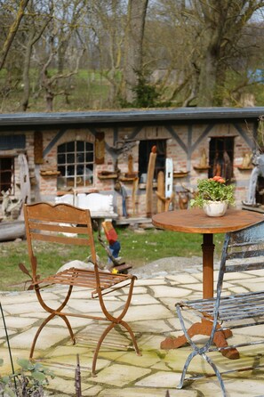 Ein rustikaler Außenbereich mit Holztisch und Metallstühlen, einem Blumentopf sowie einem Backsteinhaus im Hintergrund.