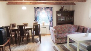 Een gezellige woonkamer met een houten plafond, een bloemenbank, een eettafel met stoelen, een televisie en een boekenkast.