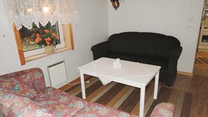 Een woonkamer met een bloemenbank, een zwarte bank, een witte tafel met opgerolde handdoeken, een radiator en een raam met kantgordijnen.