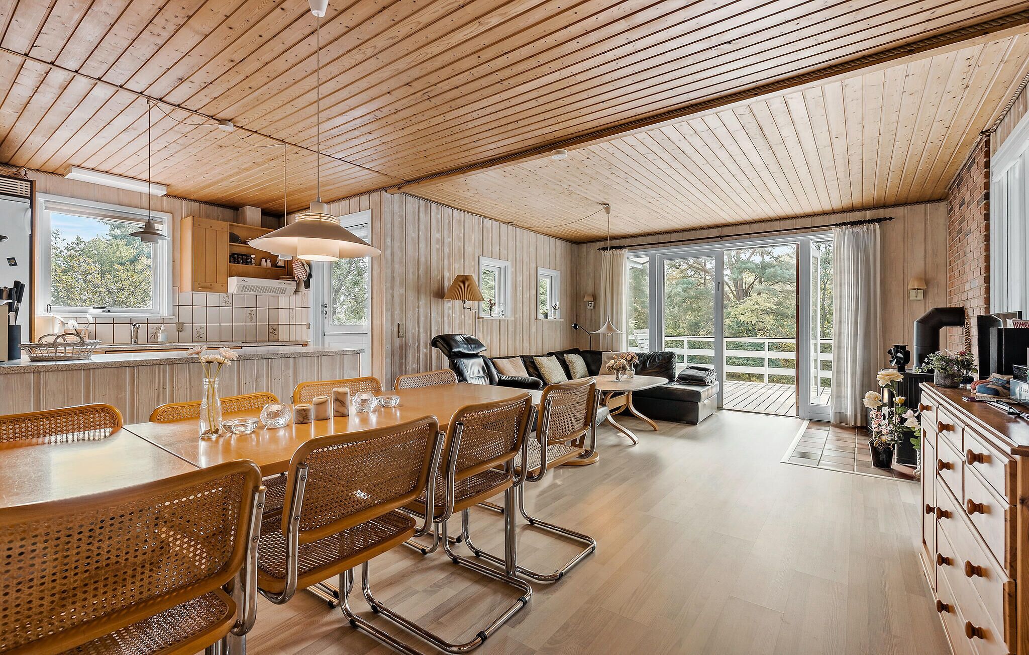 A spacious living area with a wooden ceiling, a dining table with chairs, a sofa, and a kitchen area with a window.
