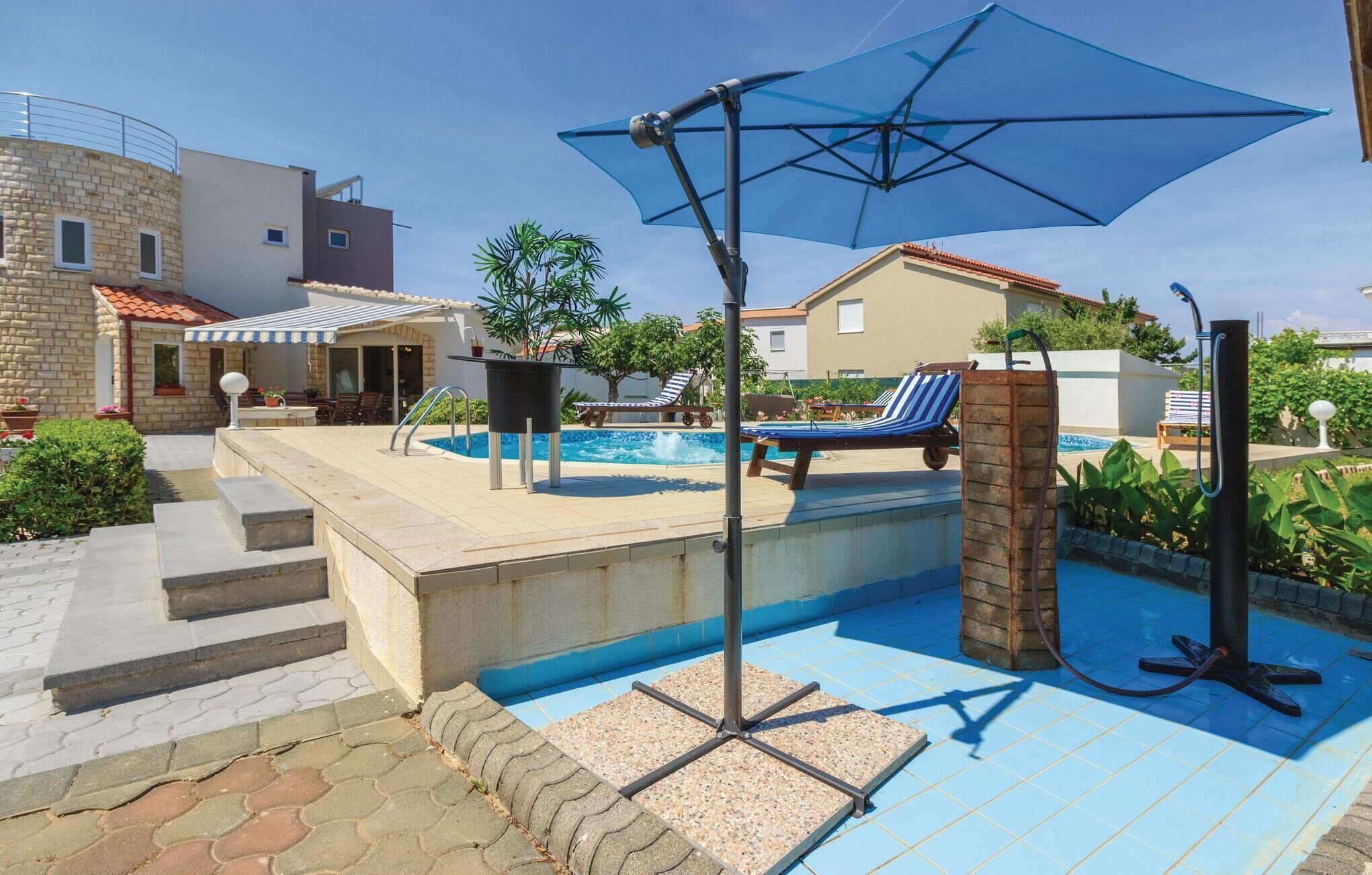 Area piscina con ombrellone blu, poltroncine da relax e una scala in pietra che conduce a un portico coperto.