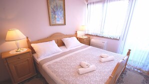 Una cama bien hecha con un edredón estampado y dos toallas enrolladas, una mesita de noche de madera con una lámpara, y un radiador en la pared.