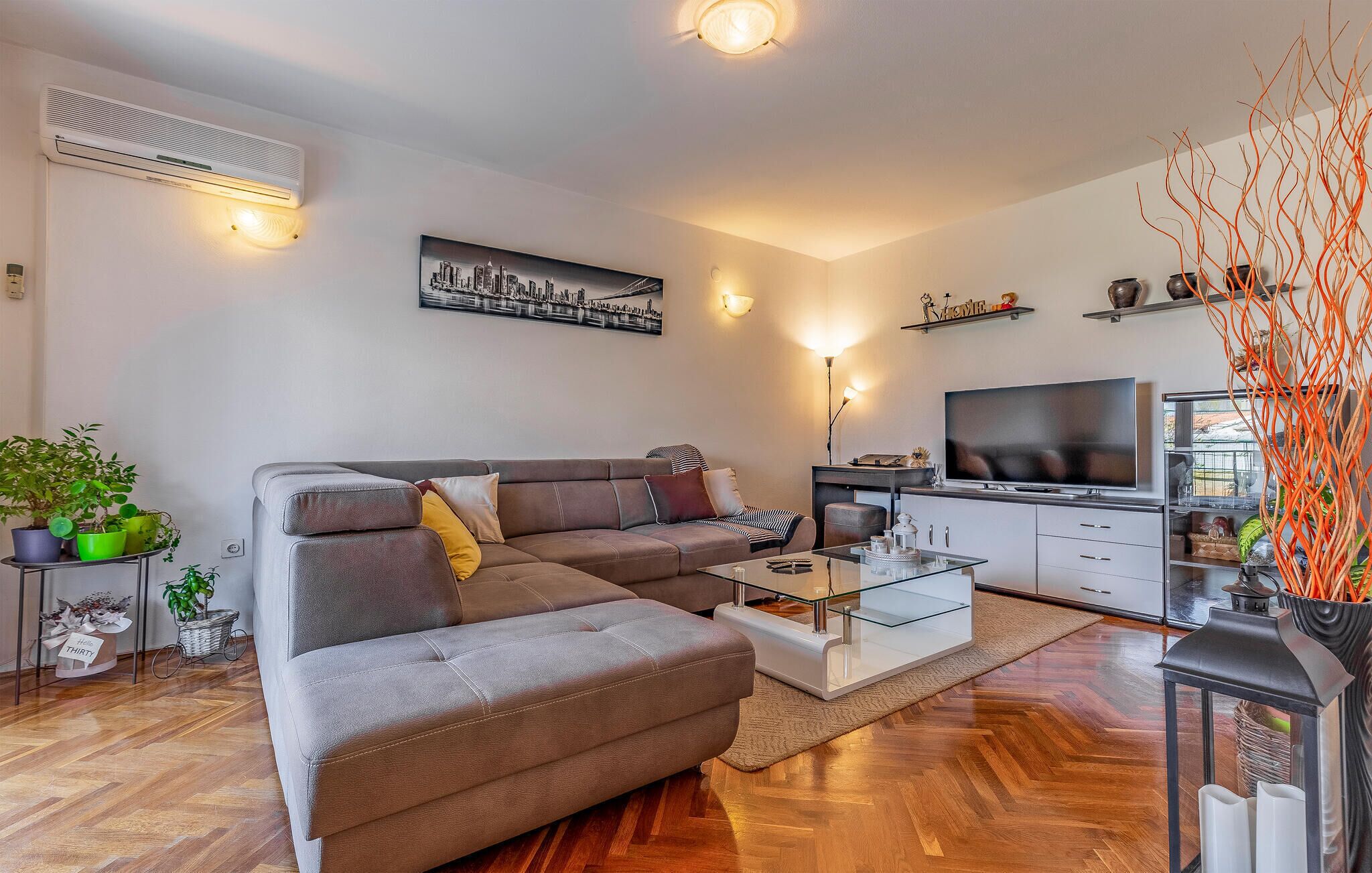 Een moderne woonkamer met een hoekbank, een glazen salontafel, een flatscreen televisie en een stadsgezicht schilderij.