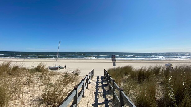 Un camino bordeando la playa que lleva al océano, con arena y algunas embarcaciones.