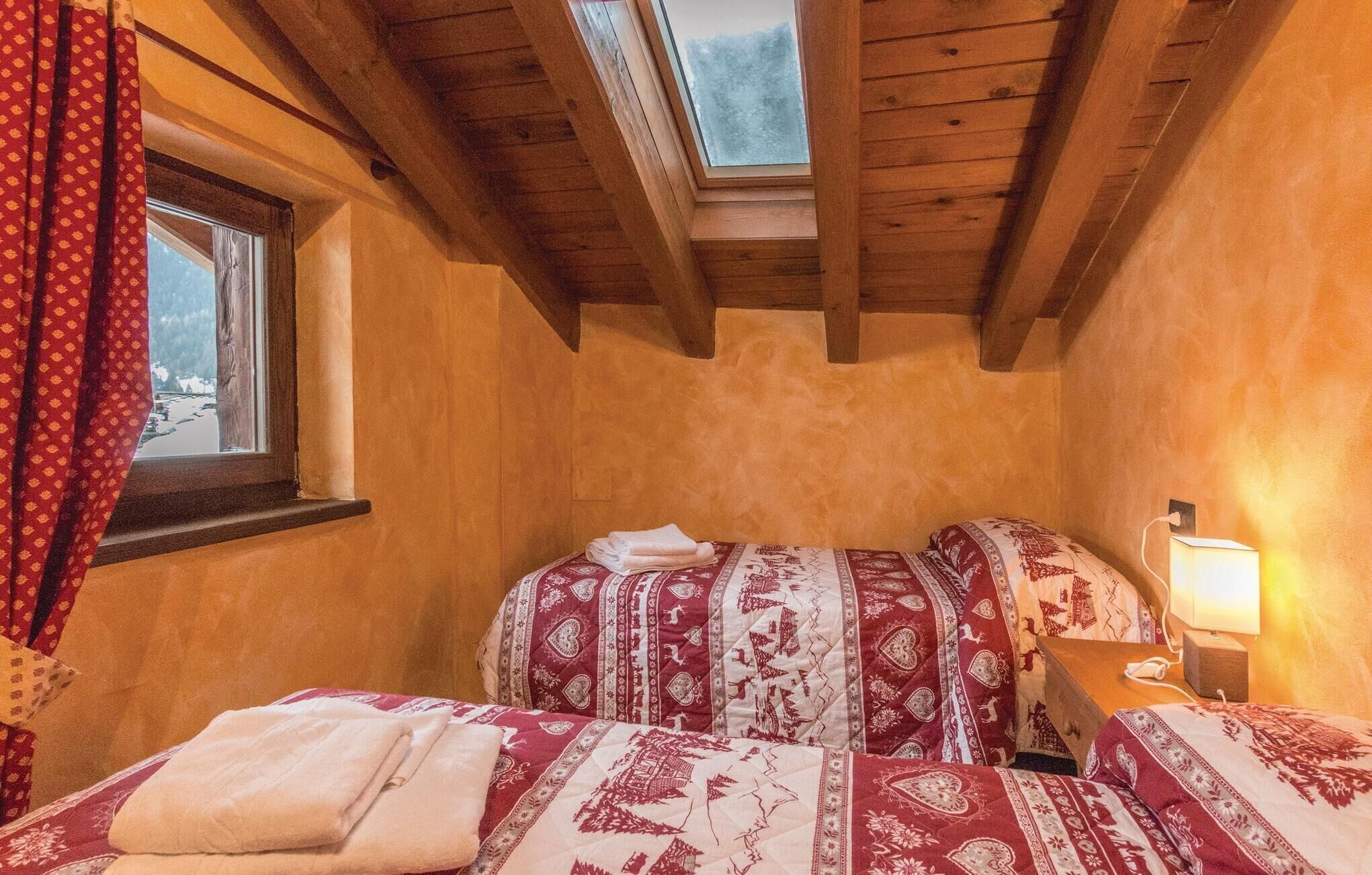 Una habitació acollidora amb sostre inclinat, un finestral, un llit amb roba de llit estampada, una tauleta de nit amb una làmpada, i una finestra amb cortines.
