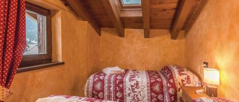 Una habitació acollidora amb sostre inclinat, un finestral, un llit amb roba de llit estampada, una tauleta de nit amb una làmpada, i una finestra amb cortines.