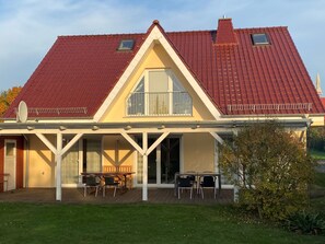 Una casa con techo de tejas rojas, un porche cubierto con mobiliario exterior y un ventanal grande.