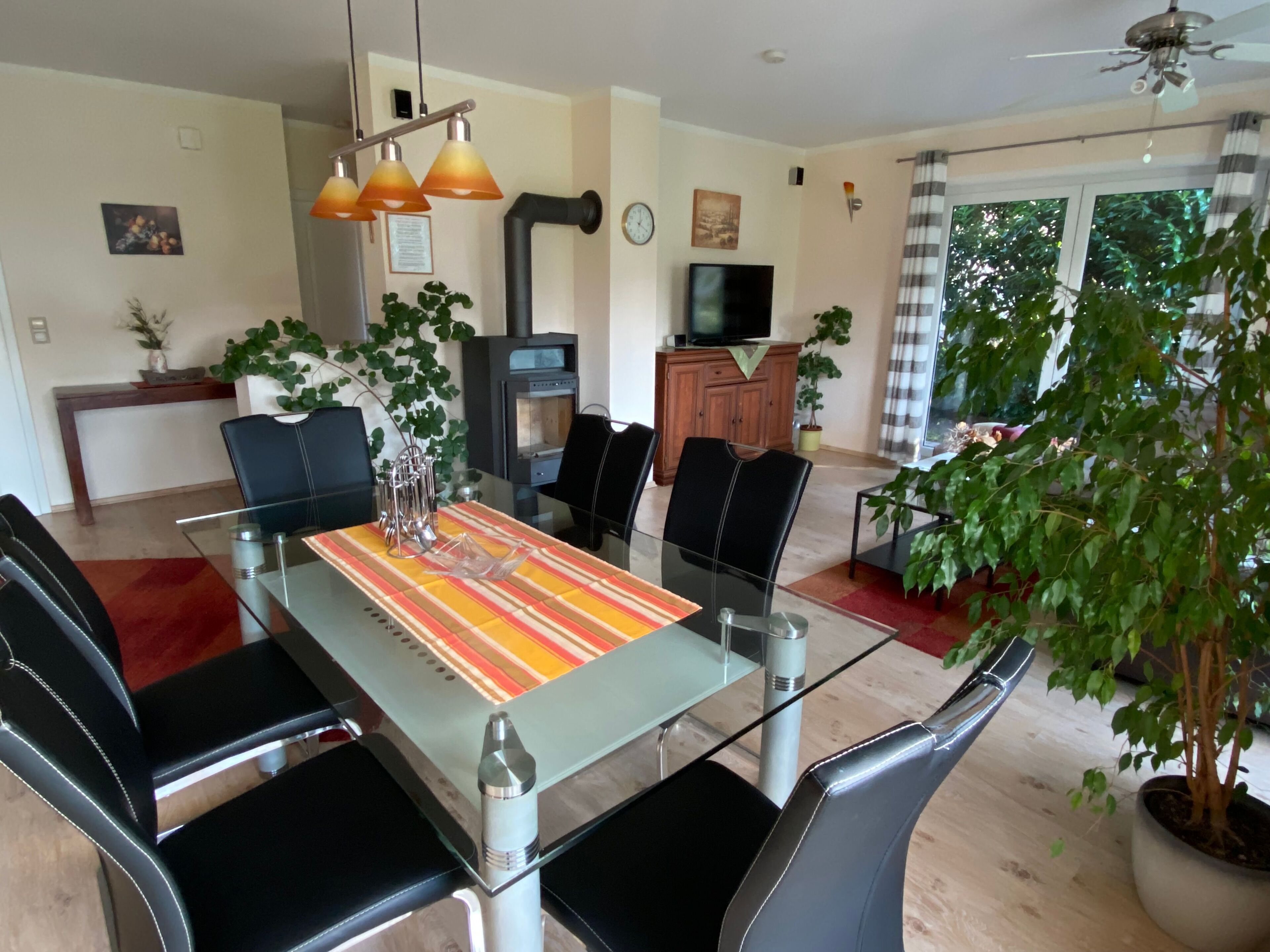 Un espace repas moderne avec une table en verre, des chaises noires et un chemin de table à rayures. On aperçoit une plante en pot sur la droite et une cheminée au fond.