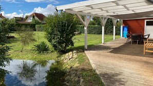 A covered outdoor area with a wooden deck, a small pond, and a garden with various plants and a tree.