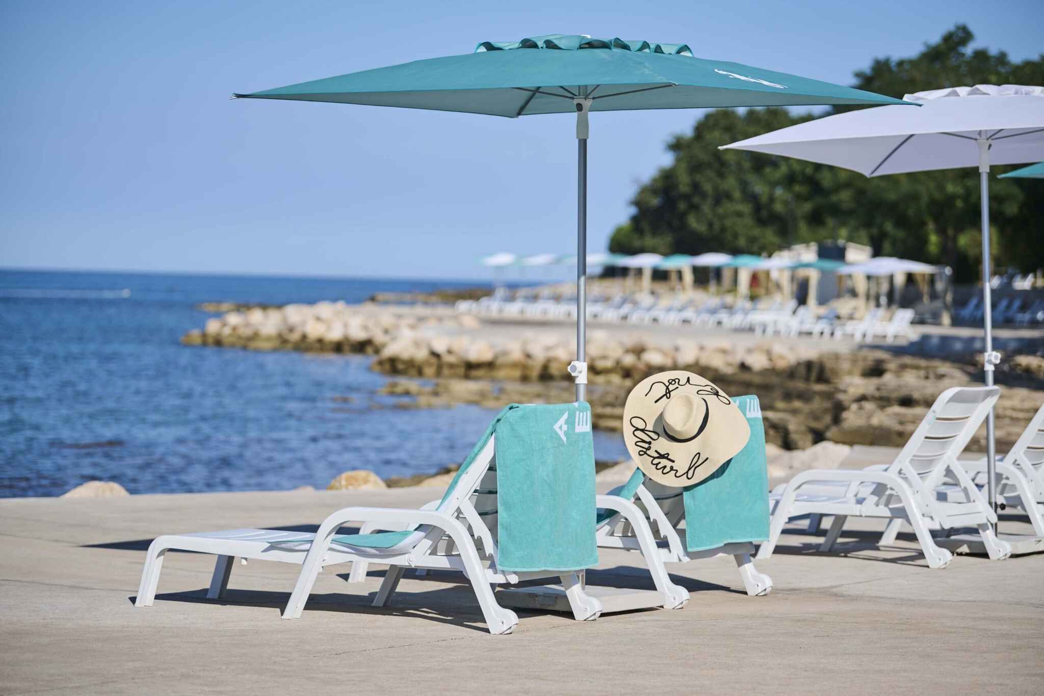 Een strandlounge met witte ligbedden en turquoise handdoeken, een bord met een hoed en zonnebril, en uitzicht op de zee en rotsachtige kustlijn.