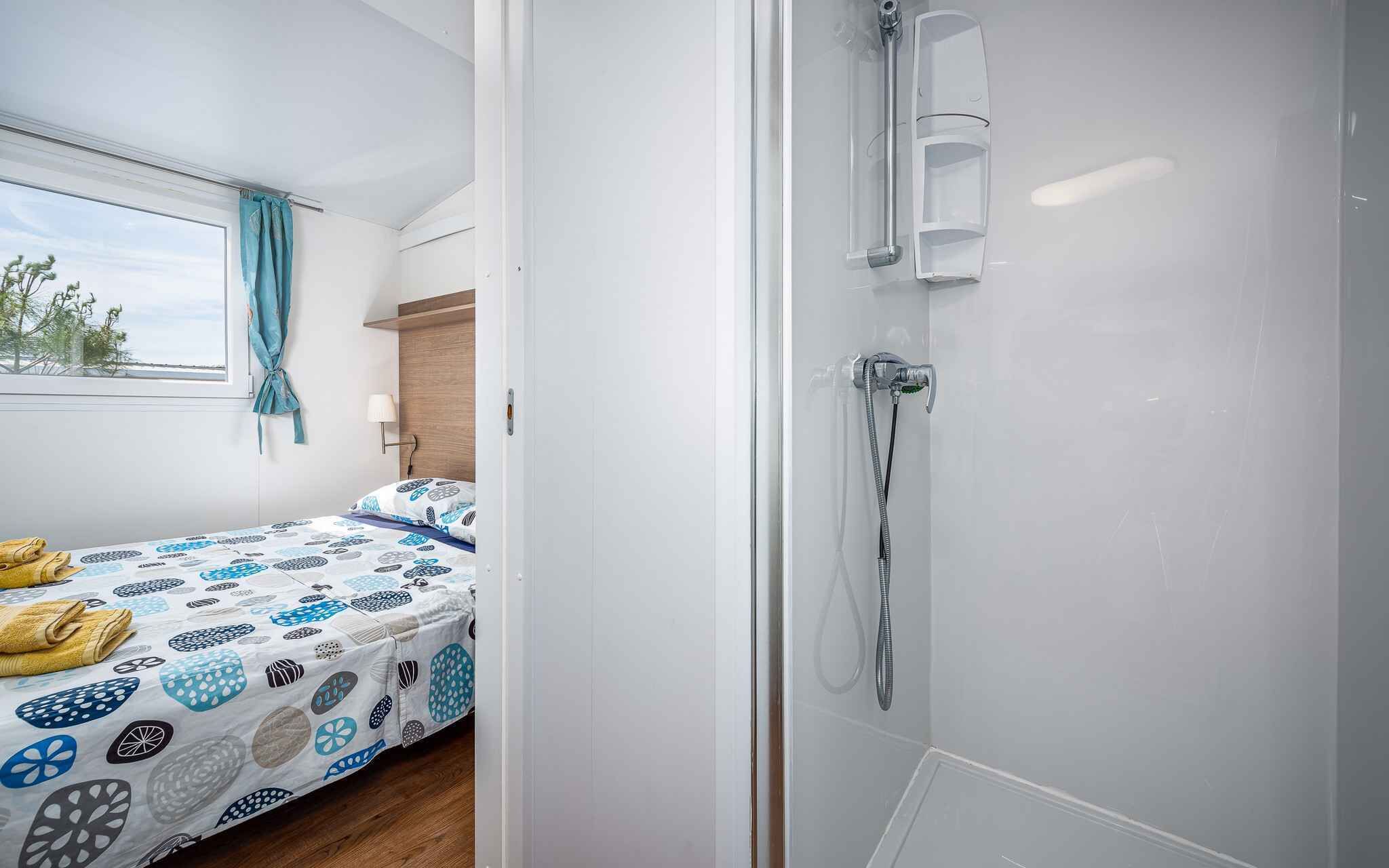 Une chambre à coucher comprenant un lit, une fenêtre avec des rideaux, une table de chevet et un espace douche doté d’une porte en verre.
