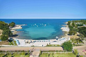 An aerial view of a coastal area with a beach, boats, and a parking lot with umbrellas.