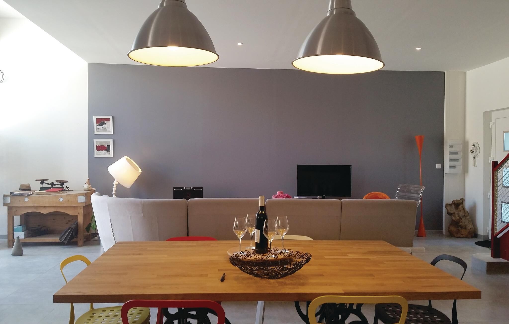A modern living room with a wooden dining table, two pendant lights, a sofa, a TV, and a small side table with a lamp.