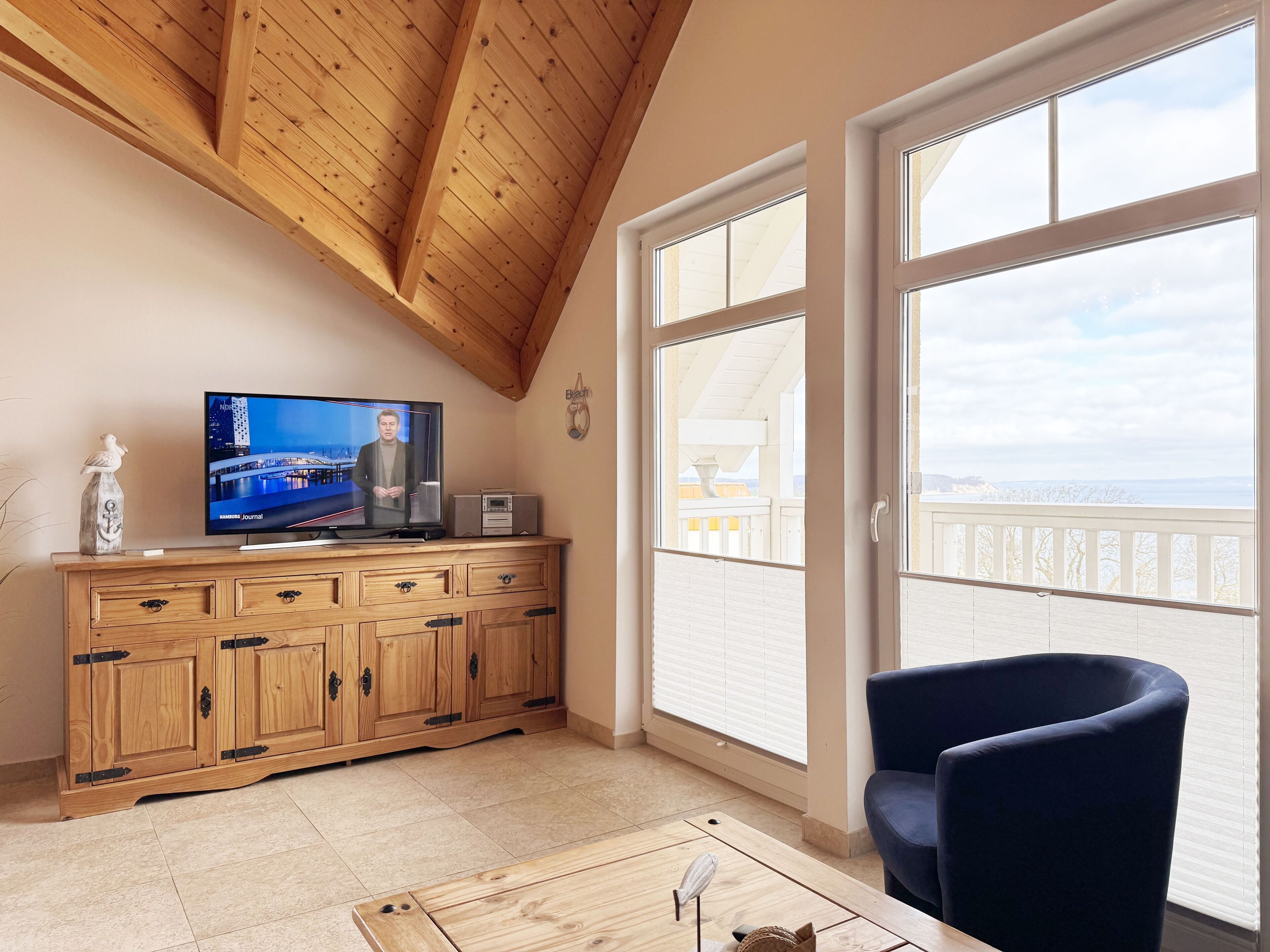 A room with a wooden ceiling, a television on a wooden cabinet, a chair, and a view of the sea through large windows.