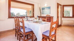 A dining area with a table set for four, wooden chairs, a large window, and a painting on the wall. - 3 bedroom cozy home in Costa Rei -CA- (Costa Rei -CA-)